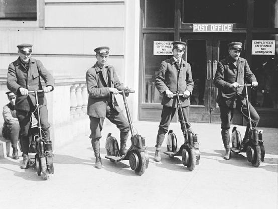 Vier Postbeamte auf eRollern vor einem Post-Office, im Jahr 1917.