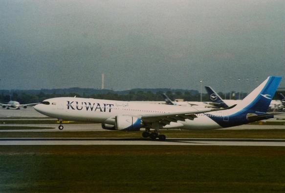 Viewed from the side, a white plane with blue details, with the word "KUWAIT" on its side, lands at an airport runway with its front wheel still in the air.
