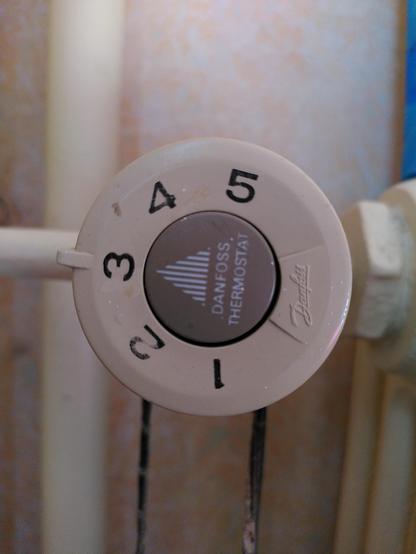 Das Bild zeigt einen weißen Thermostatknauf. Der Knauf ist ein Kreis und befindet sich vor einem weißen Heizkörper. Auf dem Knauf stehen die Zahlen 1, 3, 2, 3, 4 und 5. Um den weißen Knauf ist ein gelber Rahmen, auf dem die Wörter "DANFOSS THERMOSTAT" stehen. Der Knauf ist leicht in den weißen Heizkörper eingelassen.