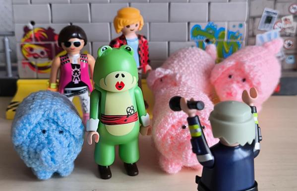Eine mit Playmobilfiguren aufgebaute Szene zeigt eine Gruppe von Demonstranten, darunter eine Person im Froschkostüm und vier haekelschweine. Vorne sieht man die Rückansicht eines Vertreters der Staatsgewalt, welcher die Arme erhebt und eine Taschenlampe in der Hand hält.