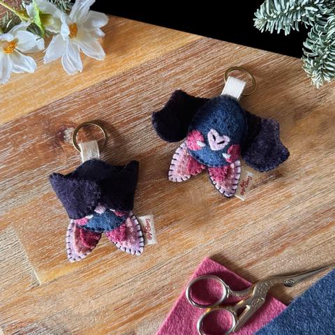 Picture of two bat keychains handmade with felt, photographed on a black and wooden background surrounded with matching felt, embroidery scissors, and a few plants.  The bats have black wings and a grey egg-shaped body, they are hanging upside down from an ivory ribbon that links them to a small bronze-coloured key ring. They have big ears made of a 3-colours gradient between dark pink and light pink. Dark pink Rosie cheeks are sewn onto either side of their faces; a little light pink heart-shaped nose is sewn between them, little nostrils are embroidered on them.  The bats on the left has its wings snapped shut with hidden magnets, revealing only two squinty eyes embroidered in white.