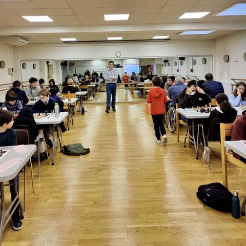 A chess tournament is taking place in a spacious room. Several tables are occupied by players, including children and adults, engaged in chess games. A person stands in the middle, possibly a referee or organizer, while a child walks past. Mirrors line one