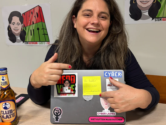 Dutch Parlement member Barbara Kathmann sits at a table holding a laptop covered in stickers related to tech and activism. A yellow sticky note on the laptop reads “Hoi Reddit!! 23-10-2025 /r/thenetherlands.” A bottle of Club-Mate is on the table, and posters with a cartoon portrait and the words “Nerd Vote” are visible on the wall behind her. Stickers on her laptop include: CYBER, Niet kutten maar patchen and All the CYBER LADIES