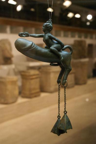 Roman bronze windchime (tintinnabulum) that depicts an erect phallus with two legs, a little erect phallus of its own and a tail that curls upward. A bell hangs on each of its little feet. A nude woman rides the phallus like a horse, extending an arm to award it a laurel wreath.