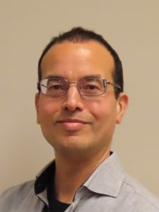 Foto van een glimlachende Eskild, korte donkere haren, bril met licht montuur, lichtkleurig overhemd