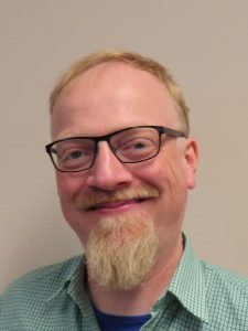 Foto van een breedlachende David, bril met donker montuur, blonde haren, snor en sikje. Groen, geruit overhemd en blauw t-shirt.