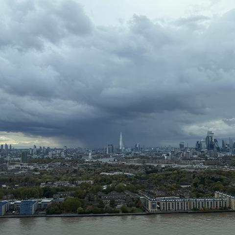 A photo of some very dark and angry looking skies over London this afternoon. Facing west, towards The Shard, we can see the Thames looking murky in the foreground. In the mid ground, we see the Shard, framed against the impressive looking clouds and what looks like some very heavy rain in the distance. Photo taken from a high-rise apartment building in Canary Wharf in the east of London. 