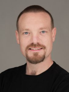 Goof kijkt vrolijk in de camera, donkerblond kort kapsel, ringbaarde en donkerkleurige trui. 