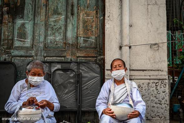 Two elderly Buddhist nuns wearing white robes and face masks pose for a portrait photo outside a weathered building in Bangkok, Thailand.