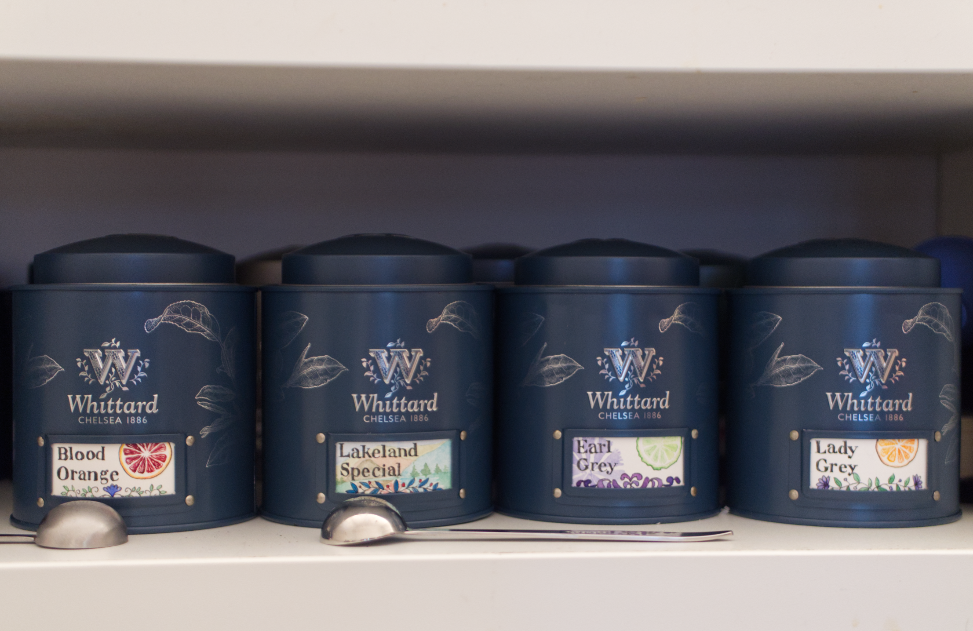 4 More stout teal tea caddies with labels that read: Blood Orange, Lakeland Special, Earl Grey, and Lady Grey.