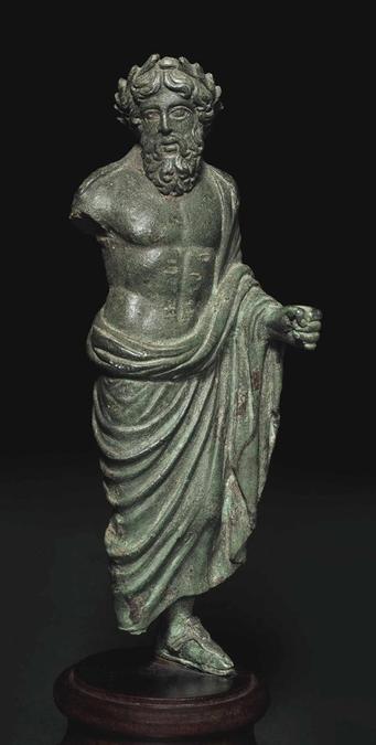 Roman bronze figurine of Jupiter as a mature, bearded man dressed in a himation that he wears around his hips and his shoulder, draped around his left arm. He is crowned with a wreath, possibly of oak leaves, his sacred plant. The right arm is, sadly, missing, as is his right foot.