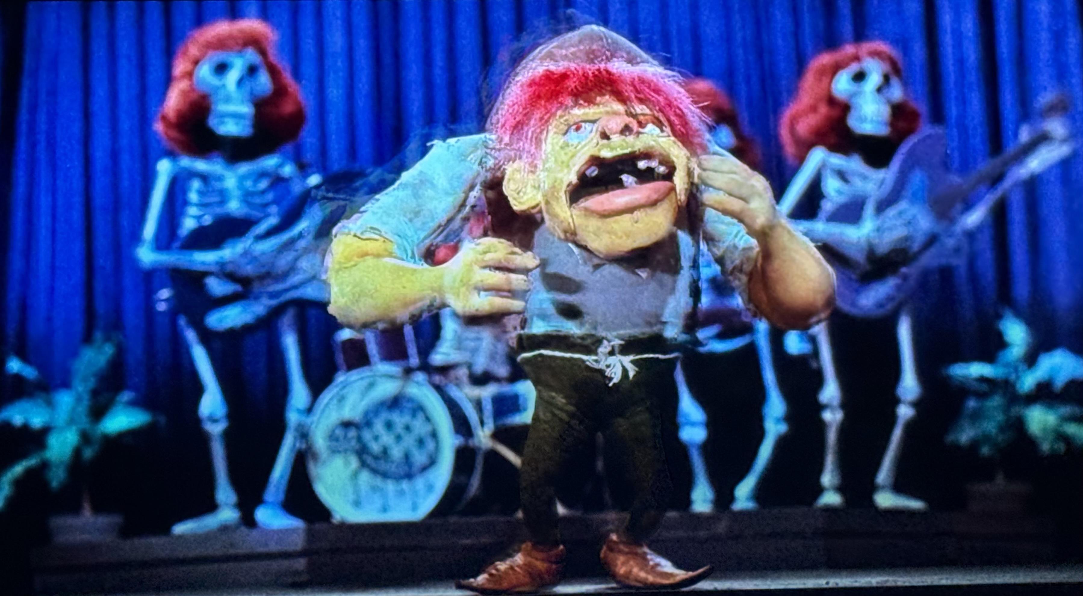 A hunchback dances in front of a skeleton band with red wigs.