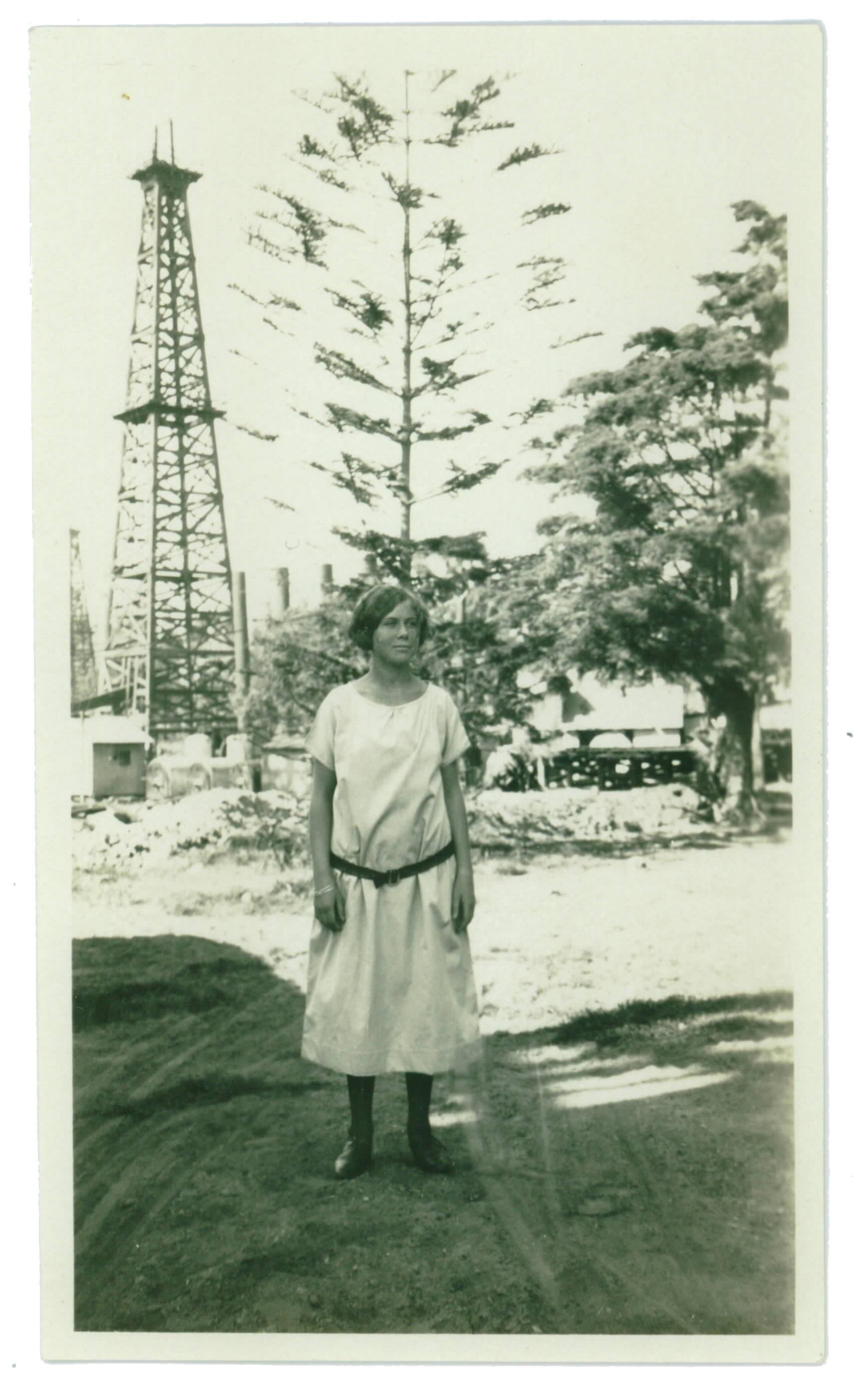Sepiatone image of a young woman in a dress, standing in front of a large oil derrick.