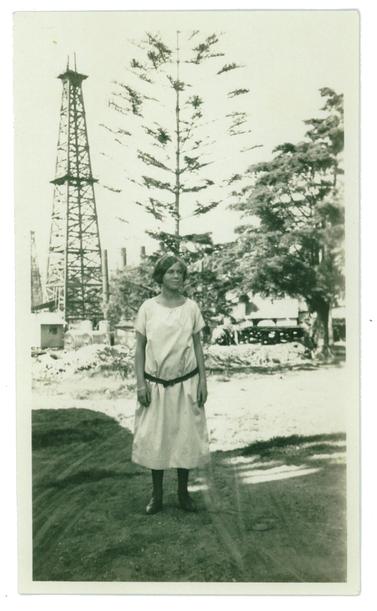 Sepiatone image of a young woman in a dress, standing in front of a large oil derrick. 
