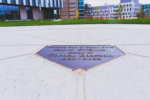 A plate in the shape of a home plate reads: Historic Site of Home Plate HOY FIELD Home of Cornell Baseball 1922-2022 in the middle of a round cement circle with grass beyond and several buildings further away