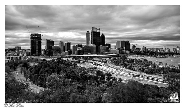 A black and white, panoramic photograph of the Perth, Western Australia central business district skyline, viewed from a high vantage point (Kings Park).
A multi lane freeway (Kwinana/Mitchell Freeways) and rail lines cut through a dense foreground of parkland and trees, leading up to the cluster of modern skyscrapers under a heavily clouded, dramatic sky.
The Swan River is visible on the far right.