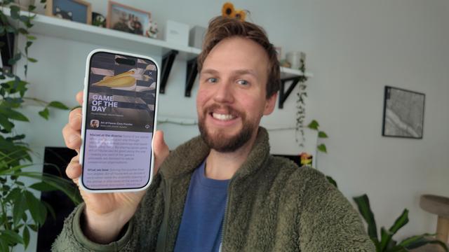 Selfie of me smiling and holding a phone displaying a "Game of the Day" screen in the App Store from Art of Fauna. Background includes shelves with plants and decor items. 