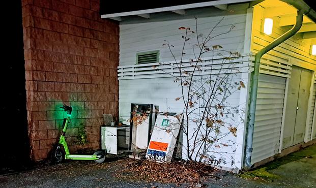 Illalla otettu kuva varasto- ja huoltorakennuksen päädystä. Seinään nojaa Teboilin piharoskis kappaleina ja muuta rojua sekä syksyisiä lehtiä. Rakennuksen kulmalla pajun tapainen hento kasvi. Rakennuksen vieressä punainen tiilimuuri, johon nojaa vihreä vuokraskootteri vikkuttaen vihreää valoa. Rakennus on valkoinen lautaverhoiltu. Rakennuksen seinässä nelionmalliset ulkovalot.