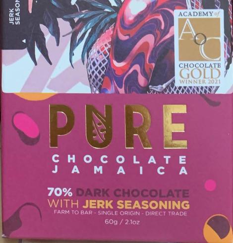 Ein Teil einer Schokoladenverpackung. Große Golde Buchstaben "PURE", darunter in weißer Schrift auf pinkem Grund „Chocolate Jamaica“ und in Weiß, Schwarz und Gelb die Sortenbezeichnung "70% Dark Chocolate with Jerk Seasoning". Oben rechts das Logo mit der Gold Auszeichnung der Academy of Chocolate.