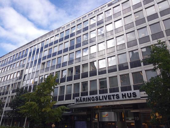 A large six storey building with many windows. A sign saying Näringslivets hus above the entrance. Two rather young and small trees in front of it. 