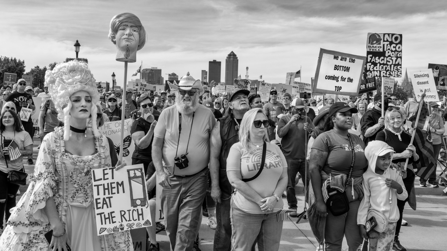 Black and white image of a No Kings Rally in Des Moines, Iowa. A woman dressed as Marie Antoinette holds a mannequin head on a stick with a sign reading, “LET THEM EAT THE RICH”.