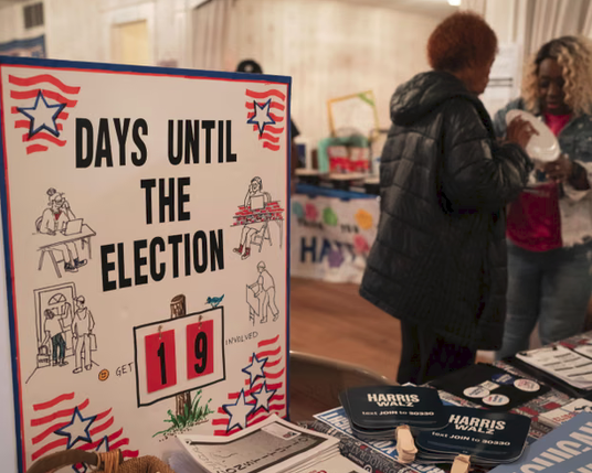 In the foreground a sign with blue stars and red site says "DAYS UNTIL THE ELECTION 19" next to a pile of Harris-Walz schwag with two black women in the background