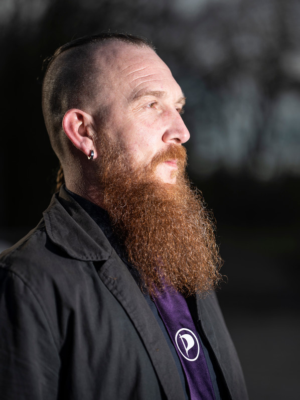 Foto van Arjan, met korte hanekam, lange rode baard, ronde oorbel zwart jasje met paarse piratenpartij stropdas op een zwart overhemd. 