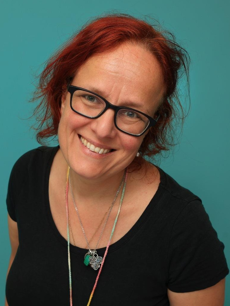 Foto van een lachende Kirsten, met halflange, rode haren, bril, kettingen op een zwart t-shirt