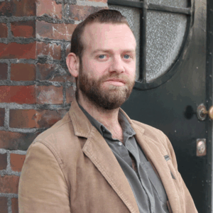 Foto van lijsttrekker Matthijs, donkerblonde haren, snor en baardje, lichtbruin jasje en groen overhemd. Op de foto leunt hij tegen een muur en is een groene deur, met rond raampje zichtbaar. 