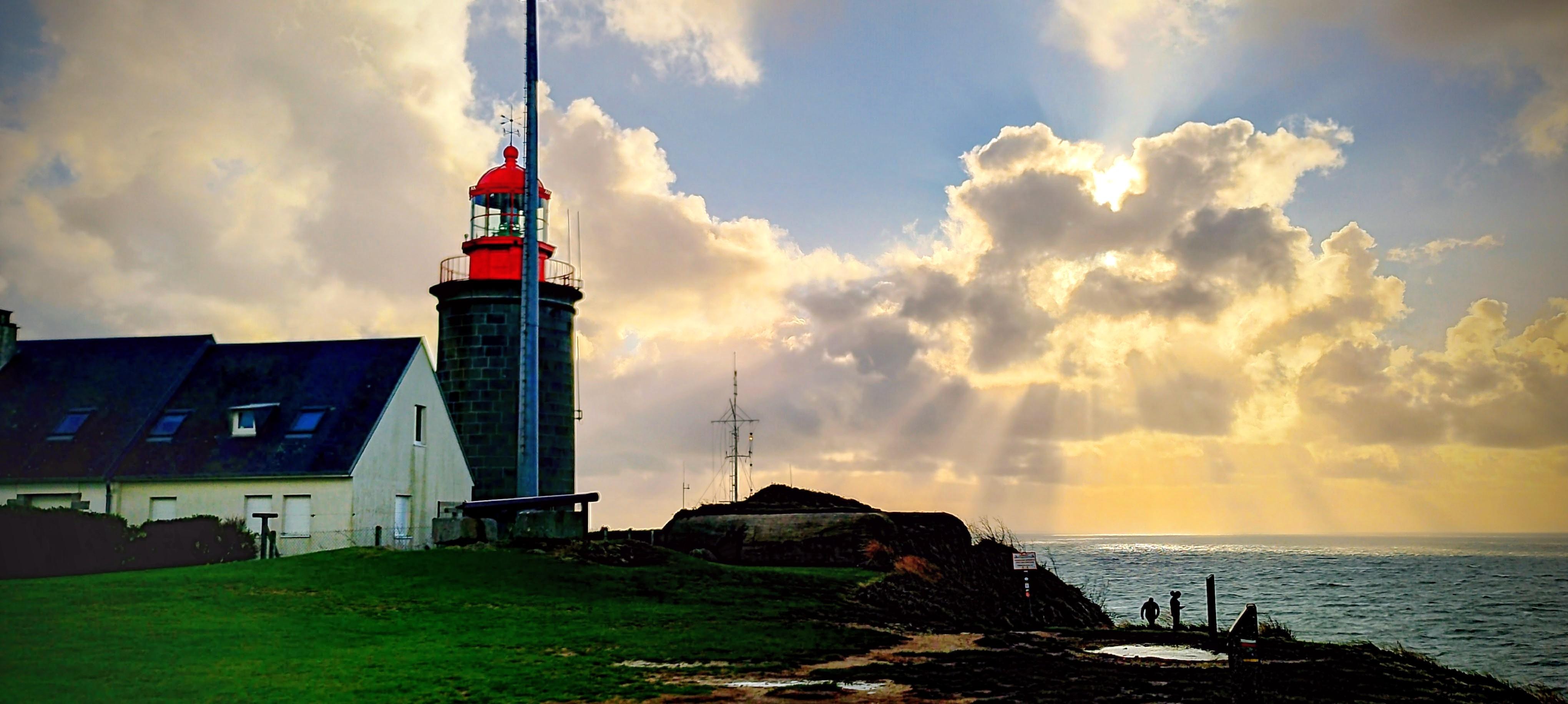 A panoramic, eye-level, wide shot shows a lighthouse complex on a grassy cliff edge overlooking the sea, under a dramatic sky where sunbeams break through clouds. On the left, a dark-colored, cylindrical lighthouse tower with a bright red lantern room stands next to a simple, white, one-and-a-half-story building with a dark roof. In the center and right, the grassy cliff extends towards the water, with a small hillock topped by an antenna structure. The sky is filled with voluminous, white and grey clouds, and on the right, bright sunbeams radiate downwards towards the choppy, dark blue sea, illuminating the horizon. Two small, silhouetted figures stand near the cliff edge on the right, looking out at the water.