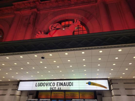 Exterior night photo of the Bill Graham Civic Auditorium marquee, showing text "Ludovico Einaudi Oct 22" in black block letters. Illuminated in red above the marquee is the fictional character Stay Puft Marshmallow Man, from the movie Ghostbusters 