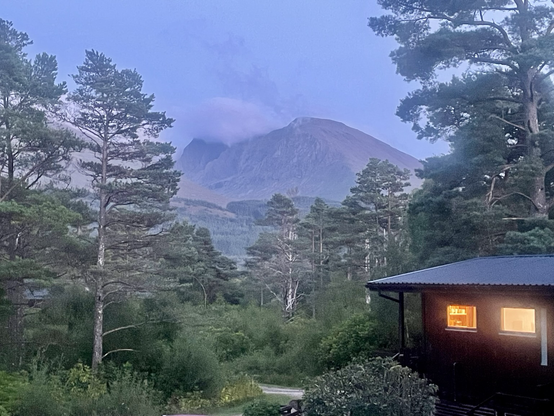 Cabin with glowing lights in the foreground. Cloud shrouded Ben Nevis in the distance, with the whole scene surrounded by large evergreen trees. 