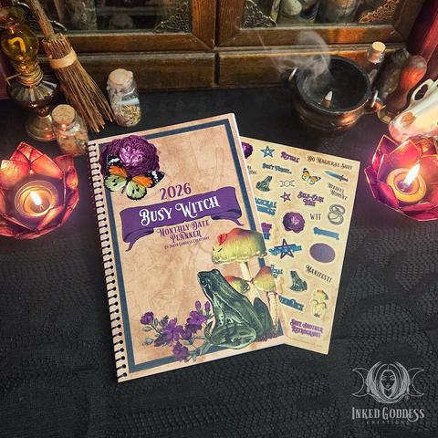 A brown spiral-bound journal lies closed on a dark lace-covered surface. The background image of the cover looks like parchment paper. It has natural imagery on it, including illustrations of a frog, flowers, a butterfly, and mushrooms. The text on the cover reads "2026 Busy Witch Monthly Date Planner." A full-color sticker sheet peeks out from under the journal that includes symbols, inspirational words, and more. This is an exclusive product from Inked Goddess Creations.