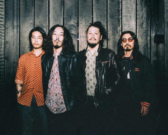 The photo features the four men of krautrock band Minami Deutsch. They are standing in close proximity to each other against a wooden backdrop, dressed casually, some are wearing leather jackets.  