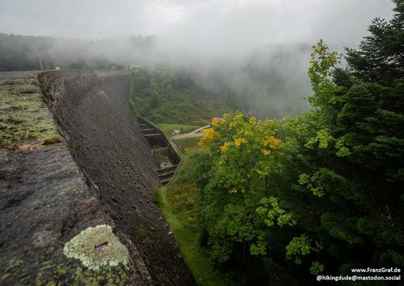 Perched high above a misty valley, this evocative scene captures the raw beauty of nature veiled in quiet mystery. A massive, weathered stone dam curves gracefully into the distance, its textured surface adorned with patches of vibrant lichen—a testament to time and the elements. The dam’s sturdy structure contrasts with the softness of the surrounding landscape, where rolling hills fade into a sea of low-hanging clouds, their edges blurred by the damp, misty air.

Below, the valley unfolds in …