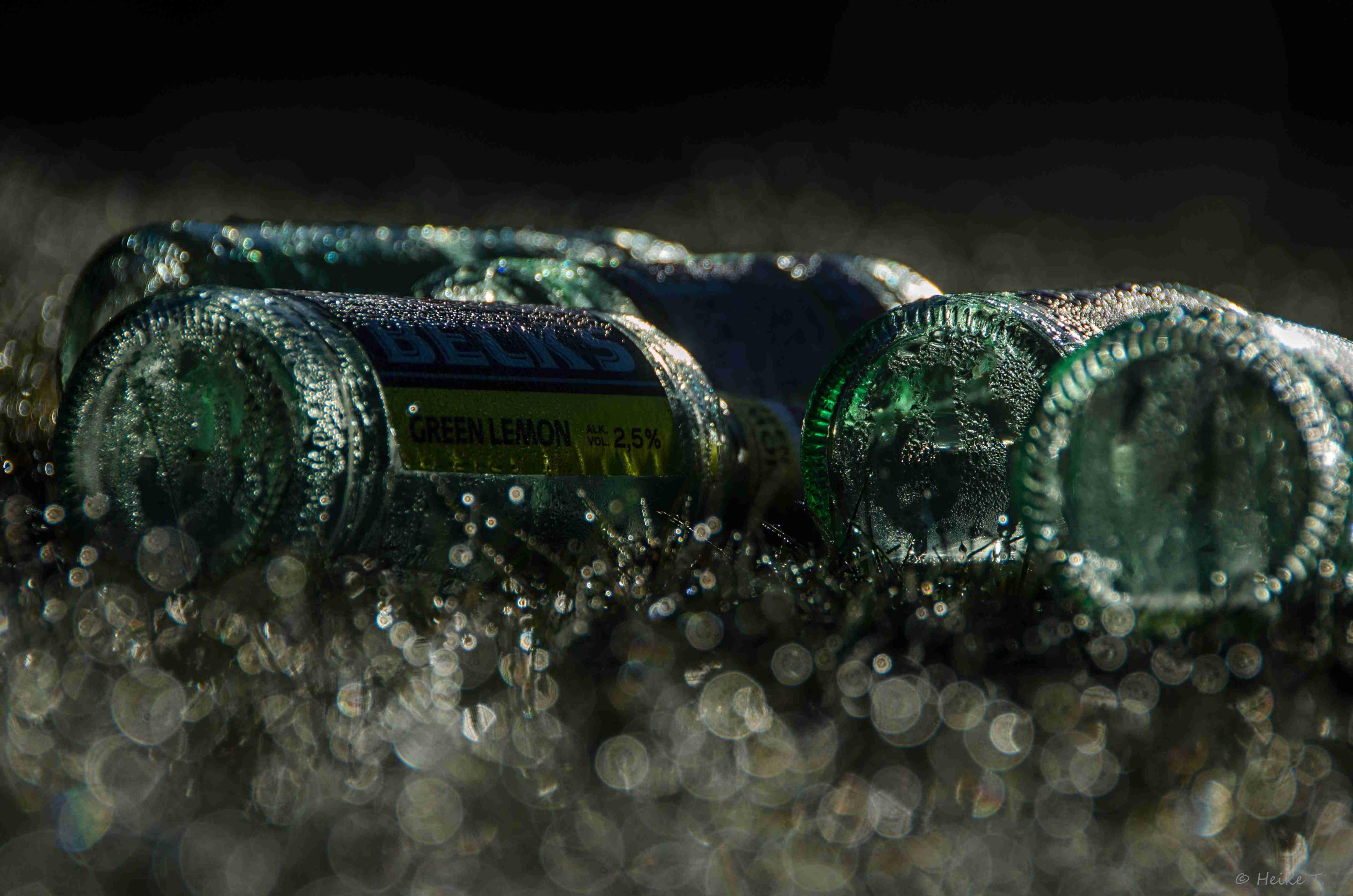 Mehrere grüne taubenetzte Flaschen,  liegend auf dunklem Wiese mit unscharfen Lichtreflexen vom Tau.