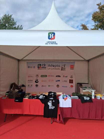 Caseta Héroes de 4 patas con su logo encima. Mesa con diferentes camisetas y merchandasing. En el fondo panel "Adopta un Jubilado" con su logo (un perro mayor en silla de ruedas con una manta) con sus colaboradoes,