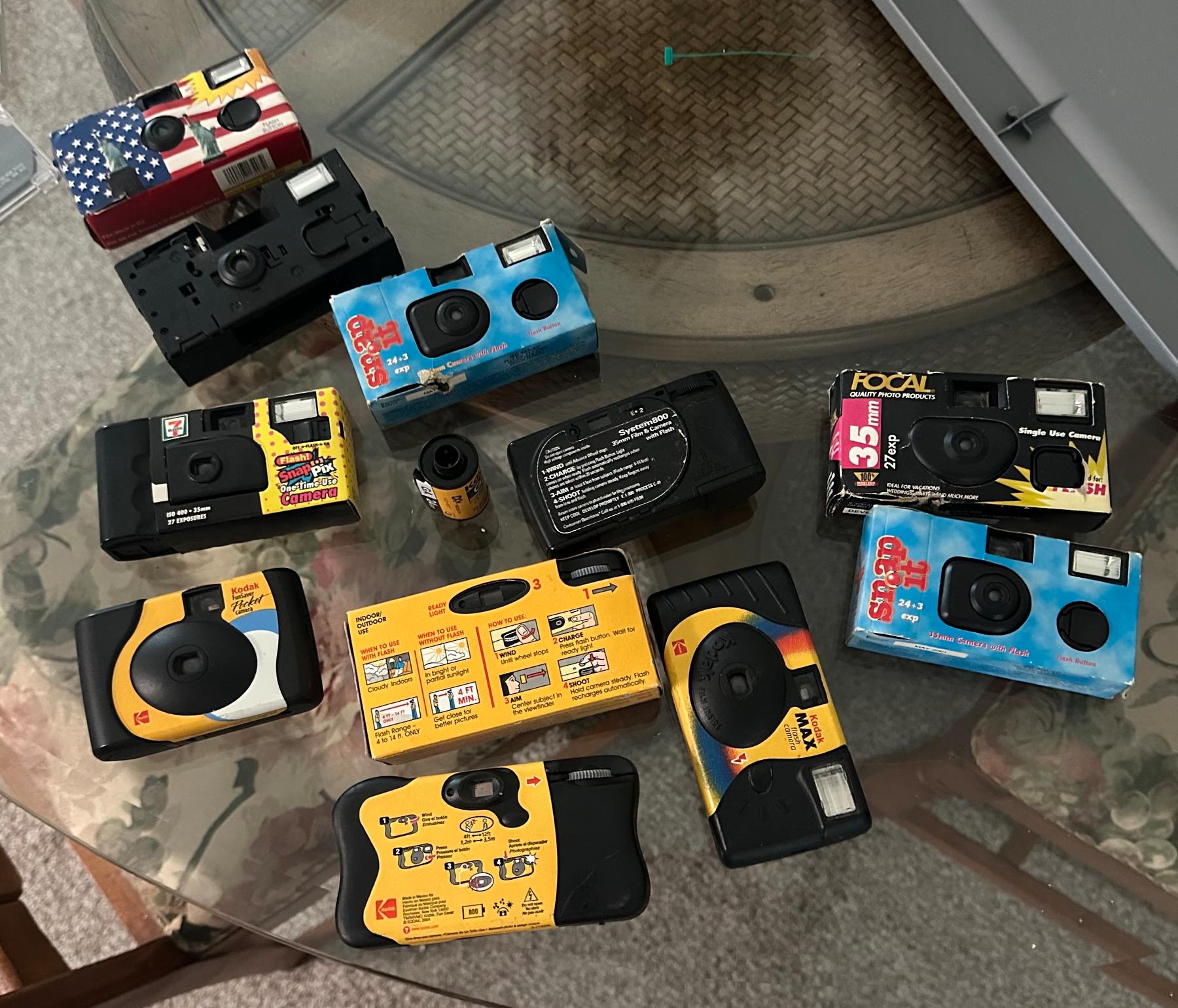 About a dozen disposable cameras laid out on a glass dinner table. 