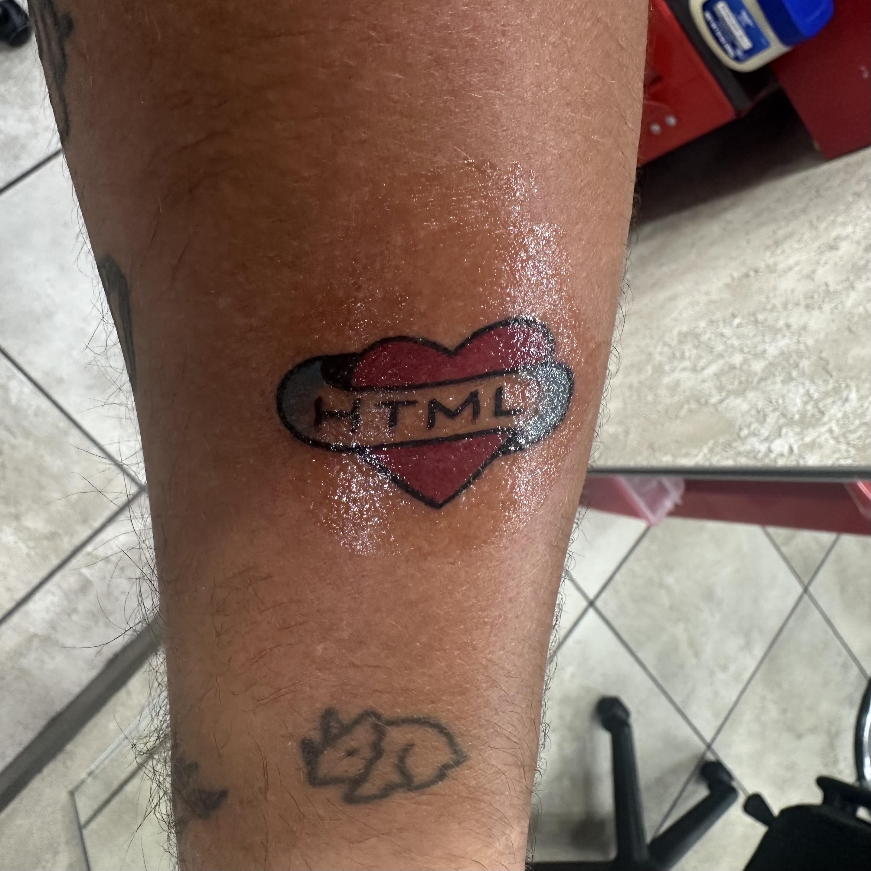 A close-up of a tattoo on a forearm featuring a heart with the text "HTML" inside it. The tattoo is located above another smaller tattoo of a dinosaur
