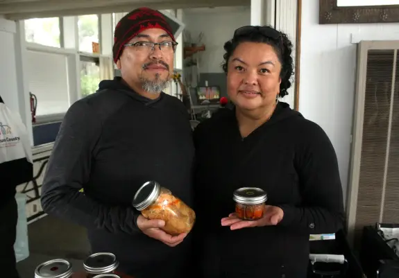 Two people standing indoors hold jars of homemade preserved foods, one large and one small, smiling toward the camera.