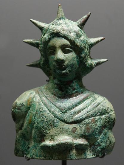 Bronze bust of Helios with a seven ray crown and long hair, wearing a chlamys cloak pinned over his right shoulder. Perhaps it is a portrait of Alexander the Great as Helios.