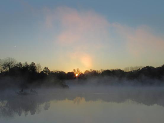 The tree lined shore of a lake, where the blue/grey mist hugging it rises into the air above and is illuminated by the glow of the sun which rises behind the trees. The sky is clear. The trees are reflected in the water. A small boat carrying two anglers drifts by on the left, it's motor slanted up out of the water.