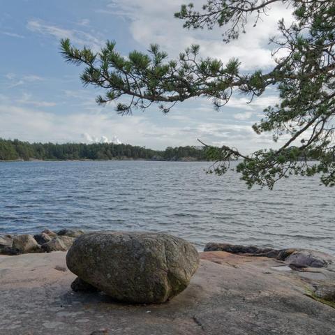 Das Bild zeigt eine Landschaft mit einem ruhigen Gewässer und felsigem Gelände. Ein großer, dunkelgrauer Felsbrocken ragt deutlich sichtbar aus einer Gruppe heller Felsen im Vordergrund hervor. Hinter den Felsen im Vordergrund erstreckt sich das Wasser bis zum Horizont und spiegelt den blauen Himmel mit vereinzelten weißen Wolken wider. Entlang des gegenüberliegenden Ufers ist eine Baumreihe zu sehen, die einen klar definierten Horizont bildet. In der oberen linken Ecke ragt ein Ast eines Nadelbaums über die Szene.