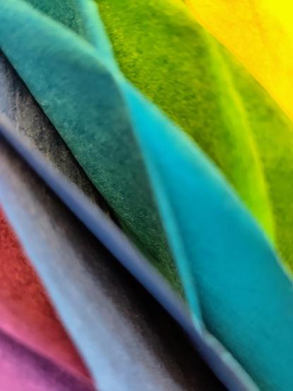 Zu sehen sind verschiedenfarbige Lamellen aus Papier. Sie geben die Regenbogenfarben wieder. Die Lamellen bilden Rauten und sind aus nächster Nähe aufgenommen. Deshalb sind die Kanten unscharf. Das wattige Papier zeichnet die Formen eher weich. Dadurch wirken die Lamellen wie gemalt. 