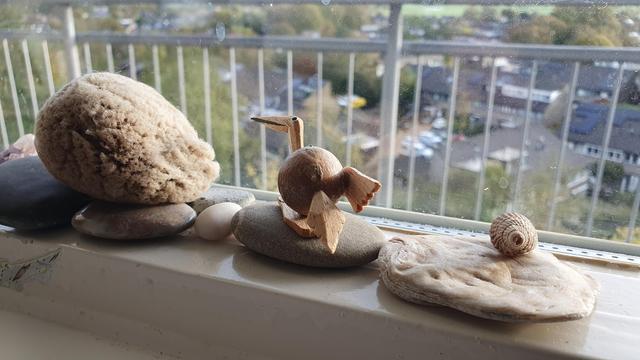 Some stones, shells and a sea sponge in a windowsill, with a small wooden bird perched on a flat stone guiding the viewer's gaze outside.