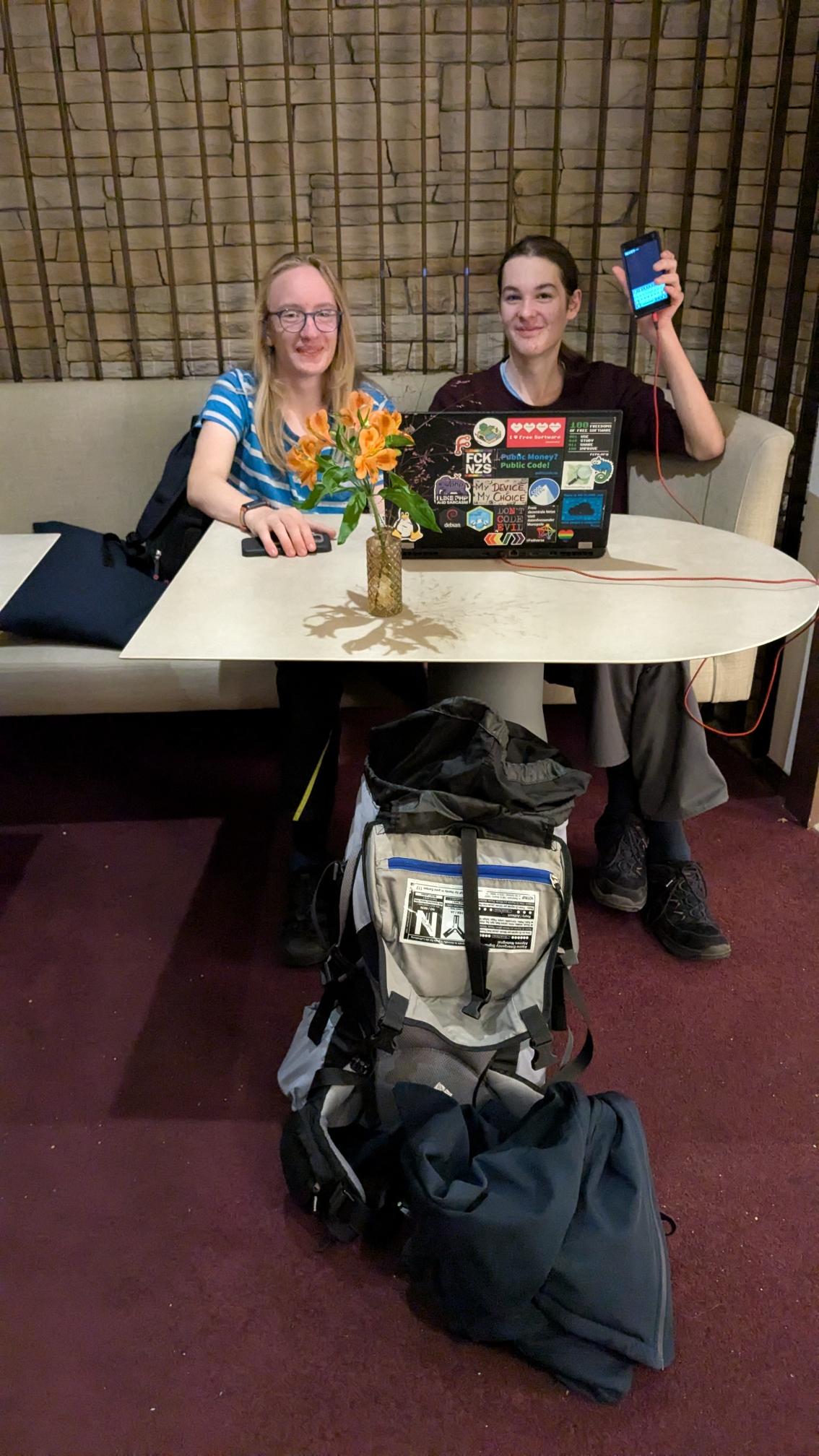 two people at a table with laptop. right person holding a phone in their hand.