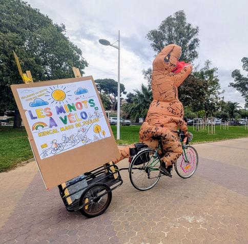 Photo of a person on a small green city bike, dressed in an inflatable T-Rex costume, pulling a small trailer holding a large poster for a bike protest type "kidical mass"