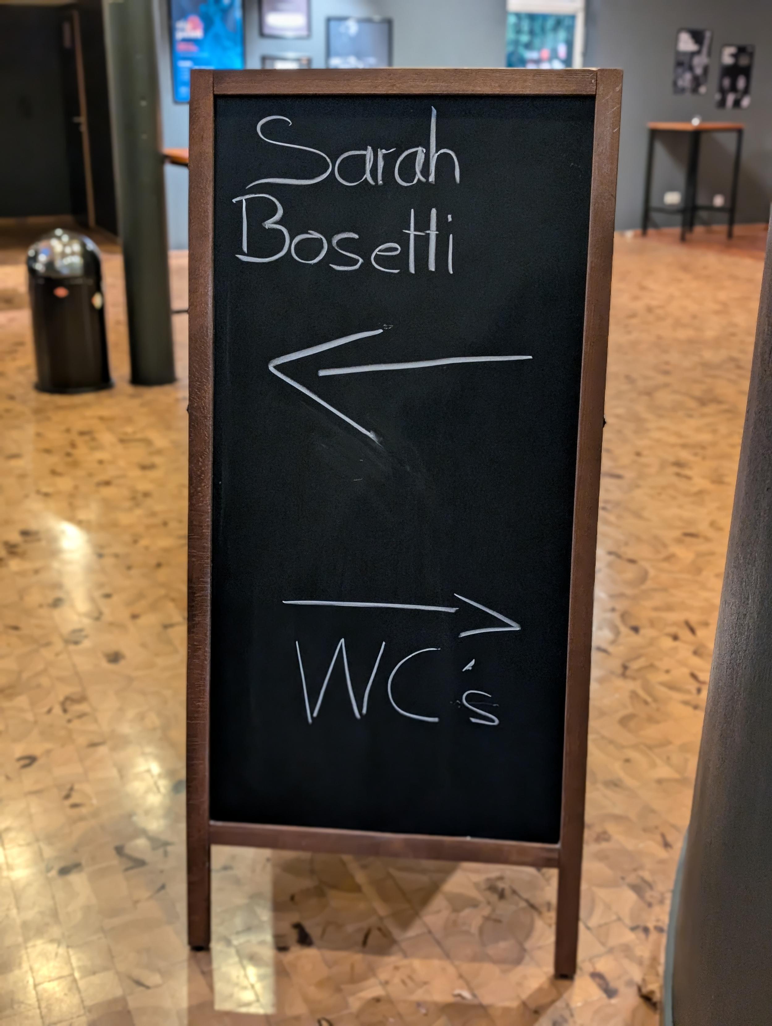 Schild: "Sarah Bosetti" mit Pfeil nach links, "WCs" mit Pfeil nach rechts