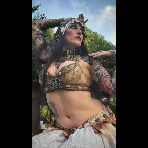 Fotografía de una mujer vestida con sujetador y falda de raqs sharqi / tribal fusion vintage, en tonos blancos y beiges, con un tocado de metal. Tiene los brazos llenos de tatuajes.