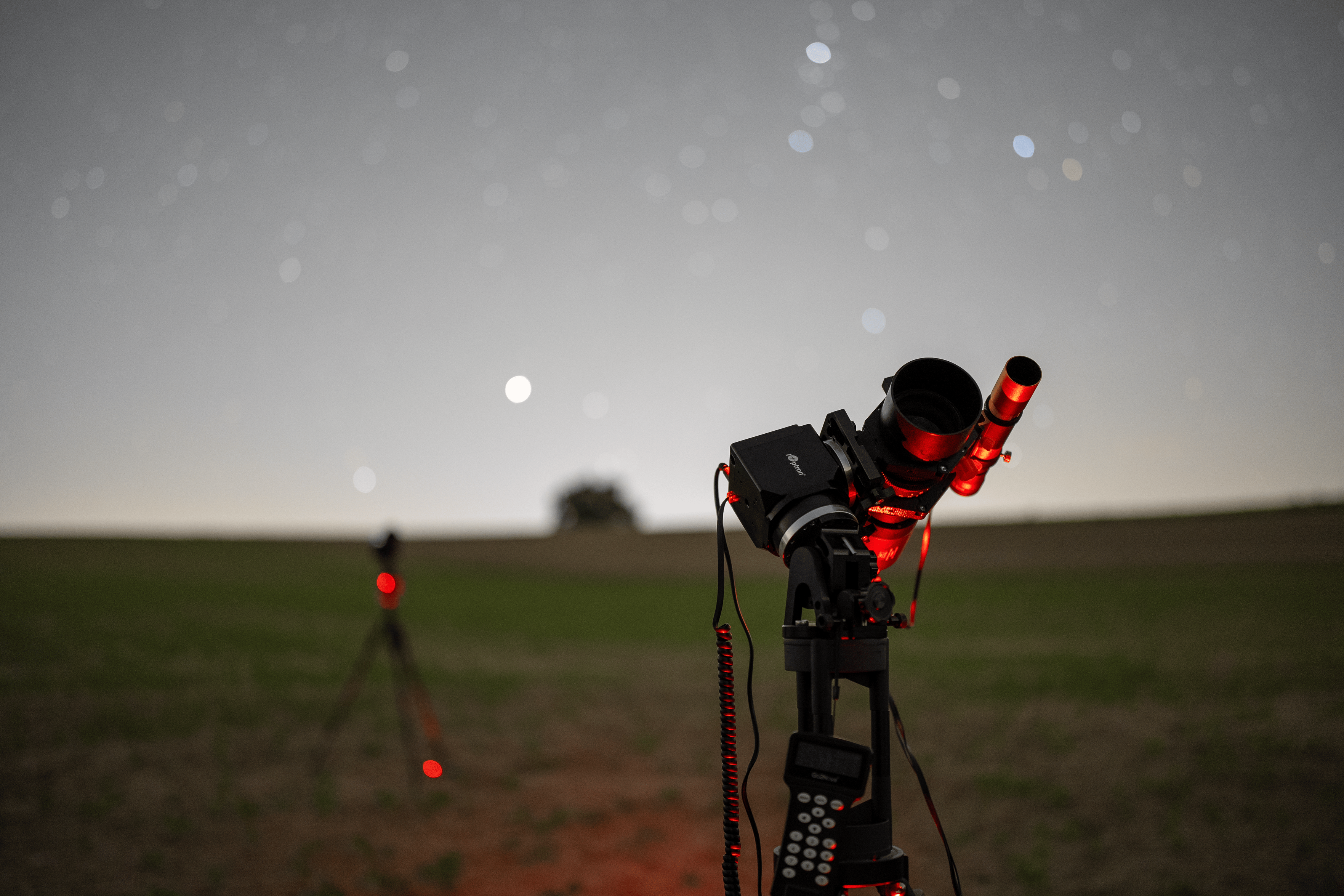 Nightscape image of a telescope setup in the center. Behind a camera on a tripod can be seen. The sky is full of stars. The scope is pointing towards the sky.